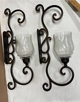 Two black wrought iron wall candle sconces with intricate curled designs and clear glass holders placed on white cloth background.