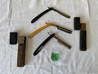 Three antique straight razors displayed with two original metal boxes on white cloth background.