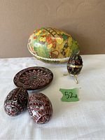 Five vintage decorative eggs and a small carved wooden plate displayed on white surface with brown background. Includes large colorful nesting egg, two dark red hand-painted eggs, one black enamel jeweled egg on stand, and small carved plate.