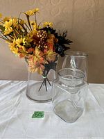 Three clear glass vases including one cylindrical vase holding artificial autumn-themed flowers with yellow daisies, orange leaves, and brown berries.