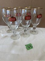 Set of six clear beer glasses with gold rims and Stella Artois logos arranged on a white cloth.