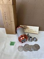 Full view of metal cookie press with box, recipe booklet and multiple cookie discs, nozzles spread around