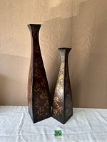 Two metal vases side by side on white cloth backdrop with beige wall background, showing square base and flared mouth with gold decorative pattern on dark metal.