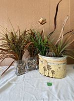 Three decorative vases with artificial plants and river rocks on a white cloth background