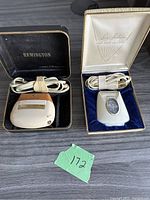 Photo showing both vintage electric razors side by side in their original cases with cords coiled inside.