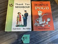 Front cover of two 1950s vintage books titled 'Thank You Neighbour' and 'The Incompleat Pogo' placed side by side on a wooden surface with lot number paper.
