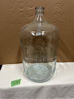 Clear glass 5 gallon jug shown upright on a white surface with a brown background. No chips or cracks visible.