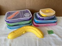 Stack of four colorful silicone collapsible containers with matching lids and attached plastic cutlery, along with a yellow banana guard placed in front.