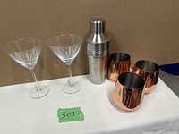 Two martini glasses, one stainless steel martini shaker, and three copper-colored metal drink cups on a table against a tan wall.