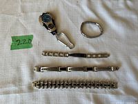 Four men's accessories including three metal bracelets and one watch keychain with carabiner clip, displayed on white cloth with measuring ruler for scale.