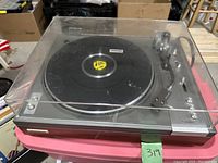 View of the turntable with closed clear dust cover on a pink surface, showing overall body and platter with 45 RPM adapter.