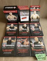 Front view of DVD cases and workout guides featuring Georges St-Pierre and the RUSHFIT branding. 9 cases arranged in 3 rows showing titles of guides and workout DVDs.