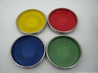 Four vintage 1970's stacking coasters showing colored fronts: yellow, red, blue, green. Metal edges with dimensional radial pattern in center.
