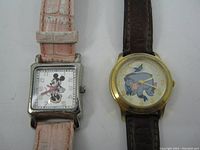 Two Disney watches placed side by side. Minnie Mouse watch has a square silver-tone case and pink strap. Eeyore watch has a round gold-tone case with a brown strap.