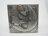 Front view of the metal artwork showing a stylized figure in relief, mounted on a square wooden base.