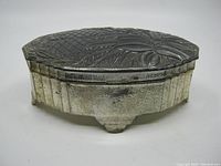 Angled side view of the oval metal trinket box showing embossed floral and geometric designs with aged patina and wear.