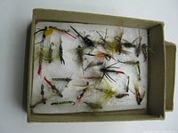 Top view of thirty assorted vintage fly fishing flies arranged in a box on white padding.