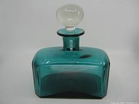 Front and slightly above angle of teal blue hand blown glass decanter with clear rounded stopper on white background.