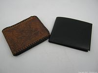 Two wallets side by side on white surface. Left is brown embossed western theme wallet with wear and leather loosening at corner. Right is new black smooth genuine leather wallet with clean appearance.