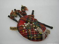 Pin cushion shaped like a Scottish bagpipe with red tartan fabric and multiple pins inserted. Shows tassel details and ribbon bow.