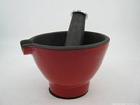 Front angled view of red enamel solid iron mortar with black interior and black iron pestle inside; shows pouring spout and round footed base.