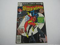 Front cover of The Spider Woman #1 comic book showing Spider Woman in red and yellow costume in an action pose, standing out against a black background with white spiderweb circle and various action elements including guns and characters in the background.