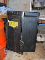 Two black plastic mechanics creepers side by side showing top surfaces and locations next to orange buckets and shelving in basement.