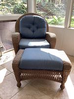 Front view of faux wicker chair with blue cushions and matching ottoman with blue cushion on tiled floor near window.