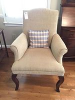 Full front view of the beige upholstered armchair showing curved wooden legs and checkered accent pillow.