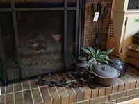 Entire fireplace area with black metal fireplace screen, fireplace tools, candle holder, cast iron pot, a metal lantern, and enamelware coffee pot arranged on the hearth. Wooden broom leaning against side wall.