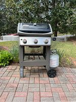 Weber propane grill on wheels with attached propane tank on bricks outdoors, showing whole unit.