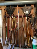Wide view of garden tools hung on wooden rack including rakes, shovels, post digger, and brooms.