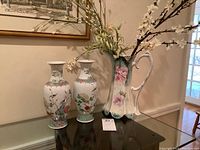 Two Asian style porcelain vases with floral and bird motifs along with a decorative tall pitcher with floral rose design and curved handle displayed on glass table.