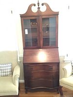 Front view of wood secretary desk with two glass doors, two shelves inside, drop-front writing surface, and three drawers at bottom with ornate handles.