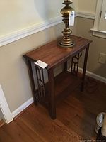 Wooden side table with a brass lamp placed on top, showing the front and left side against a wall with baseboard molding.