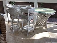 Photo showing the set of three white wicker plant stands and one round wicker table with glass top placed on a tiled screened porch.