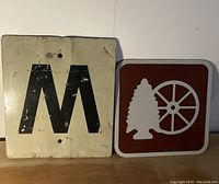 Two metal signs shown side by side on a surface against a wall, close view of the large sign with the black 'M' on an off-white background and another brown sign with white tree and wheel graphics.