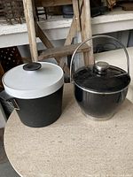 Both ice buckets shown together on a surface; one black with white lid and the other glossy black with metal handle.