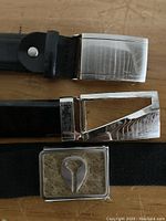 Close-up of three belt buckles including a silver rectangular buckle with a keyhole shape, a plain brushed metal buckle, and a shine silver buckle with classic design