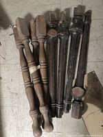 Two sets of eight vintage wooden table legs, four are lighter wood and four are dark stained wood, laying on a tile floor, showing overall condition and design