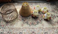 Photo shows vintage floral-pattern rug with a beige background and floral motifs in pink, green, and cream. On top of the rug are five lampshades: two large cone-shaped woven cane lampshades, three small cream-colored lampshades decorated with molded flower accents in pink, yellow, and beige.