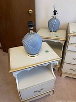 Two white night stands topped with blue ribbed ceramic lamps without shades. Night stands have gold trim and single decorative drawer pulls. Light wear on edges visible.
