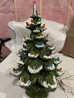 Front view of the green ceramic Christmas tree showing artificial snow and multicolored plastic lights with electric cord visible.