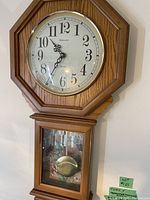 Octagonal wooden pendulum wall clock with white face, black numerals, black decorative hands, and glass panel showing pendulum