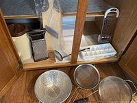 Kitchen cabinet holding various kitchen utensils including a plastic container, two metal box graters, wooden cutting board, metal ladle, metal colander, two fine mesh strainers, and two white plastic ice trays.