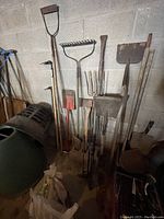 Photo of various large metal garden tools with wooden and metal handles stacked upright against a concrete block wall including rakes, shovels, spades, and forks.