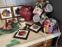 Overall view of the vintage kitchenware set including large leaf platter, red apple container, mugs, jars, framed tiles, and bucket with mugs.