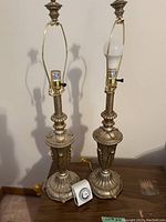 Two antique brass finish table lamps with intricate detailing and ornate base. One lamp has a bulb installed. A white timer switch is in front of the lamps. Yellow cords attached.