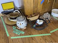 Photo of the entire lot showing owl cookie jar, woven baskets, ceramic duck pitcher, wire basket tray, trivets, glass covered butter dish, and casserole dish.