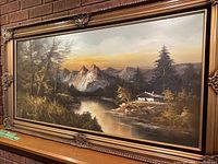 Frontal view of the framed oil painting depicting a mountain landscape with river and cabin, signature visible at lower left.